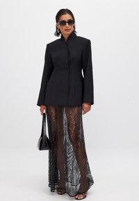 Musta strukturoitu bleiseri korkean kauluksen kanssa, yhdistettynä läpikuultavaan mustaan hameeseen, jossa on aaltoileva kuvio, pitäen pientä mustaa laukkua.