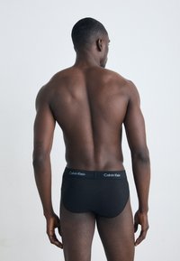 Schwarze Baumwollslips mit einem logodatierten Bund, figurbetontem Design, glatter Textur und tiefem Schnitt, von hinten betrachtet.