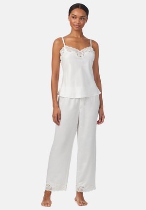 Mujer con camisola de satén blanca con borde de encaje y pantalones de pierna ancha a juego, de pie descalza frente a un fondo liso.