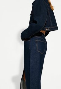 Tmavě modrá džínová bunda s krátkým střihem, se zlatými knoflíky a kontrastním stehem. V kombinaci s odpovídajícími džíny s vysokým pasem.