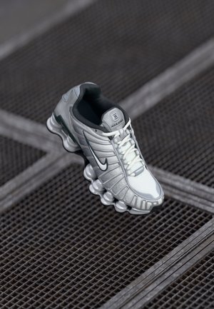 Baskets Nike Shox grises et blanches avec logo swoosh noir, lacets blancs et semelle visible de style ressort sur une grille métallique.