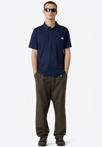 Granatowa koszulka polo z krótkim rękawem i kieszenią, zestawiona z ciemnozielonymi spodniami. Model ma na sobie okulary przeciwsłoneczne i brązowe buty turystyczne.