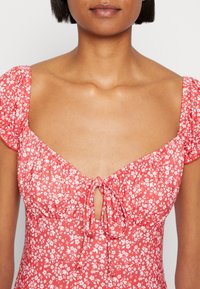 Czerwona sukienka w kwiaty z krótkimi bufkami, zaciągnięta w okolicy dekoltu z detalem kokardy, wykonana z lekkiego materiału o gładkiej fakturze.