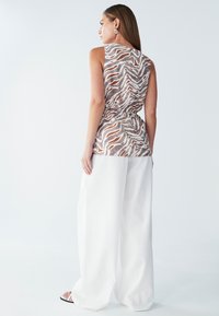 Ærmeløs top med et lyst brunt og hvidt bølgemønster, samlet ved taljen. Kombineret med løstsiddende hvide bukser.