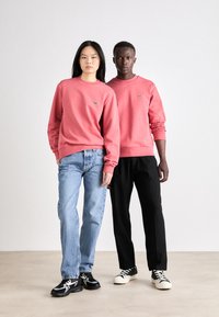 Két személy azonos rózsaszín pulcsit visel, amelyen egy logó található. Az egyik világoskék farmert, a másik fekete nadrágot és sportcipőt hord. Egyszínű háttér.