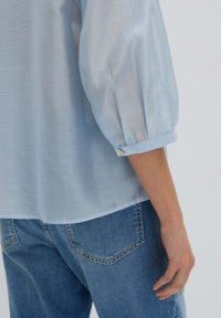Blusa azzurro chiaro, semi-trasparente, con maniche a sbuffo, polsini arrotolati e una texture liscia; abbinata a jeans in denim blu classici.