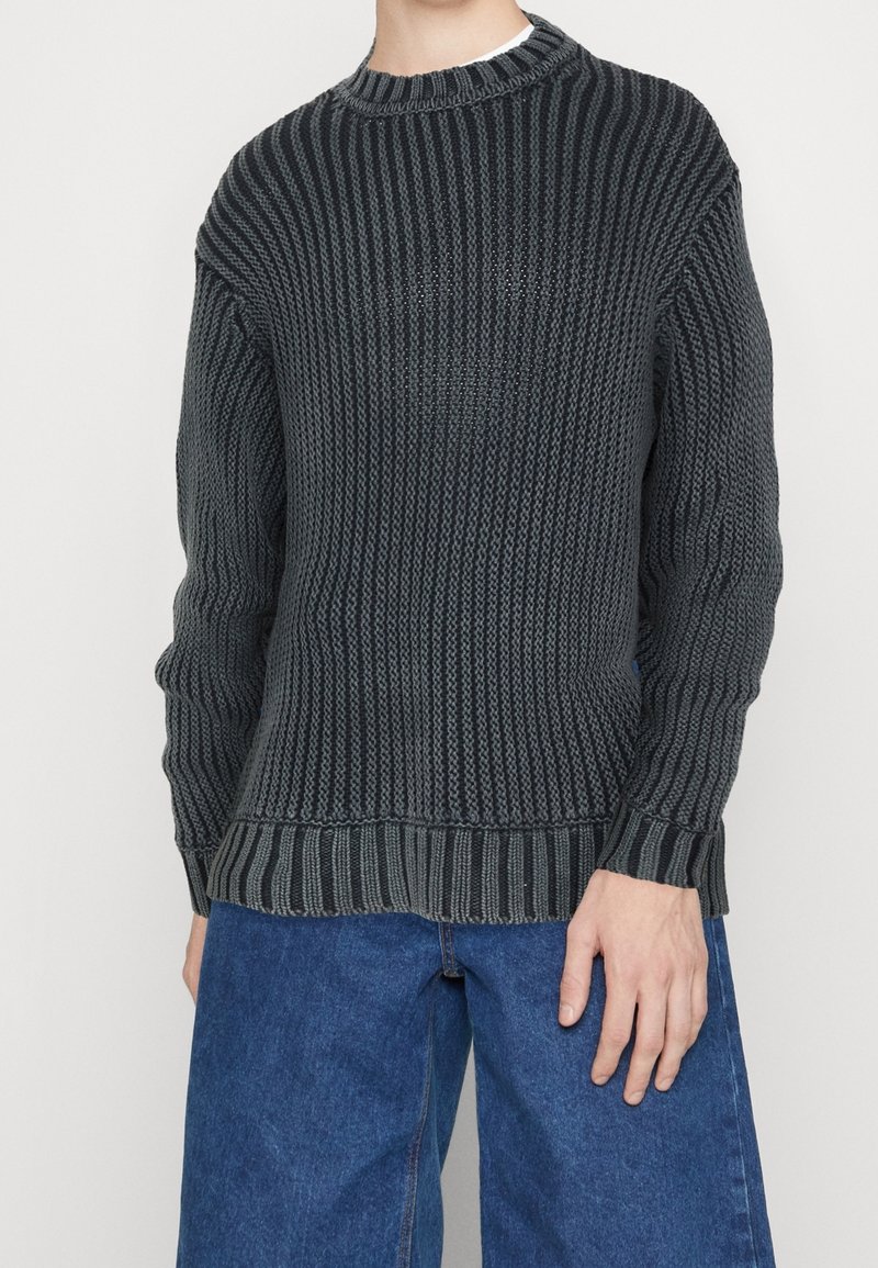 Pull en maille côtelée gris foncé porté avec un jean denim bleu ample sur une personne debout devant un fond blanc.