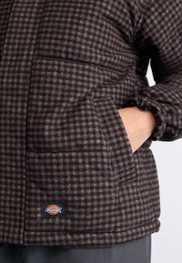 Veste matelassée à carreaux bruns avec une poche zippée, des poignets élastiques et un patch logo. Le tissu a un aspect doux et texturé.