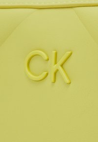 Geltona dirbtinės odos medžiaga su lygiu paviršiumi, su įspaustu "CK" logotipu atitinkamos spalvos. Nusagstytas dizainas pabrėžia paviršių.