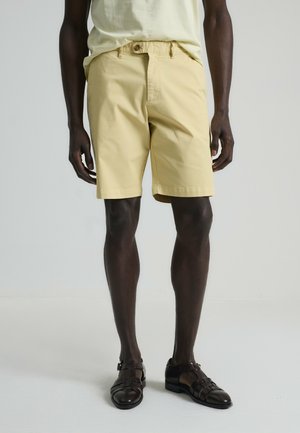 Bermudas de algodón amarillo claro con un corte relajado, que presentan un cierre de botón y cordón ajustable. Combinadas con zapatos de piel trenzada marrón oscuro.