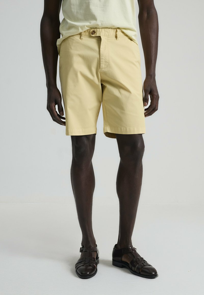 Bermudas de algodón amarillo claro con un corte relajado, que presentan un cierre de botón y cordón ajustable. Combinadas con zapatos de piel trenzada marrón oscuro.