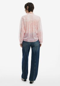 Blusa leggera rosa chiaro, trasparente, con maniche lunghe e scollatura arricciata, abbinata a jeans in denim blu a gamba larga con cuciture a contrasto.