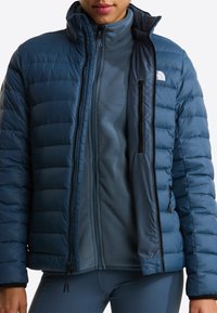 Person wearing a blue quilted jacket open over a blue fleece zip-up and blue leggings, showing layered cold-weather clothing.