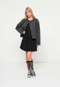 Γκρι κοντό σακάκι με στρογγυλή λαιμόκοψη, πάνω από μαύρο πουκάμισο με κουμπιά και μαύρη φούστα που φαρδαίνει προς τα κάτω, συνδυασμένα με μαύρα αθλητικά παπούτσια και κάλτσες μέχρι το γόνατο.