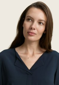 Dunkelblaue Bluse mit V-Ausschnitt und subtilen Nahtdetails an der Vorderseite. Glattes Material mit leichter Textur.