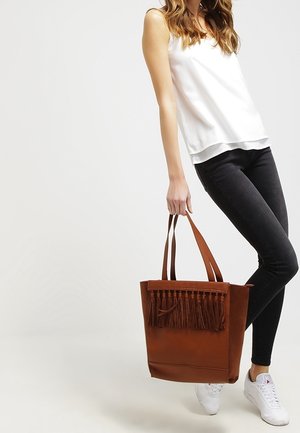 Brun läderväska med fransdetaljer upptill, platt botten och två axelband; hålls av en person som bär en vit topp och svarta byxor.