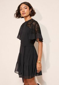 Juoda suknelė su nėrinių viršumi, trumpomis žaismingomis rankovėmis, sluoksniuotu raukinio sijonu ir surišama juosmens dalimi. Lygus, lengvas audinys su laisvai krentančiu dizainu.