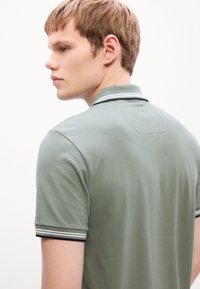 Ung mand med kort lyst brunt hår iført en grøn polo-shirt med sort og hvid stribet krave og ærmekanter, ansigtet vendt mod venstre.
