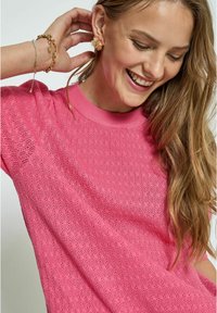 Rosa Strickpullover mit strukturiertem Muster, ausgestattet mit kurzen Ärmeln und einem gerippten Rundhalsausschnitt. Die Accessoires umfassen goldene Blumenohrringe und ein Armband.