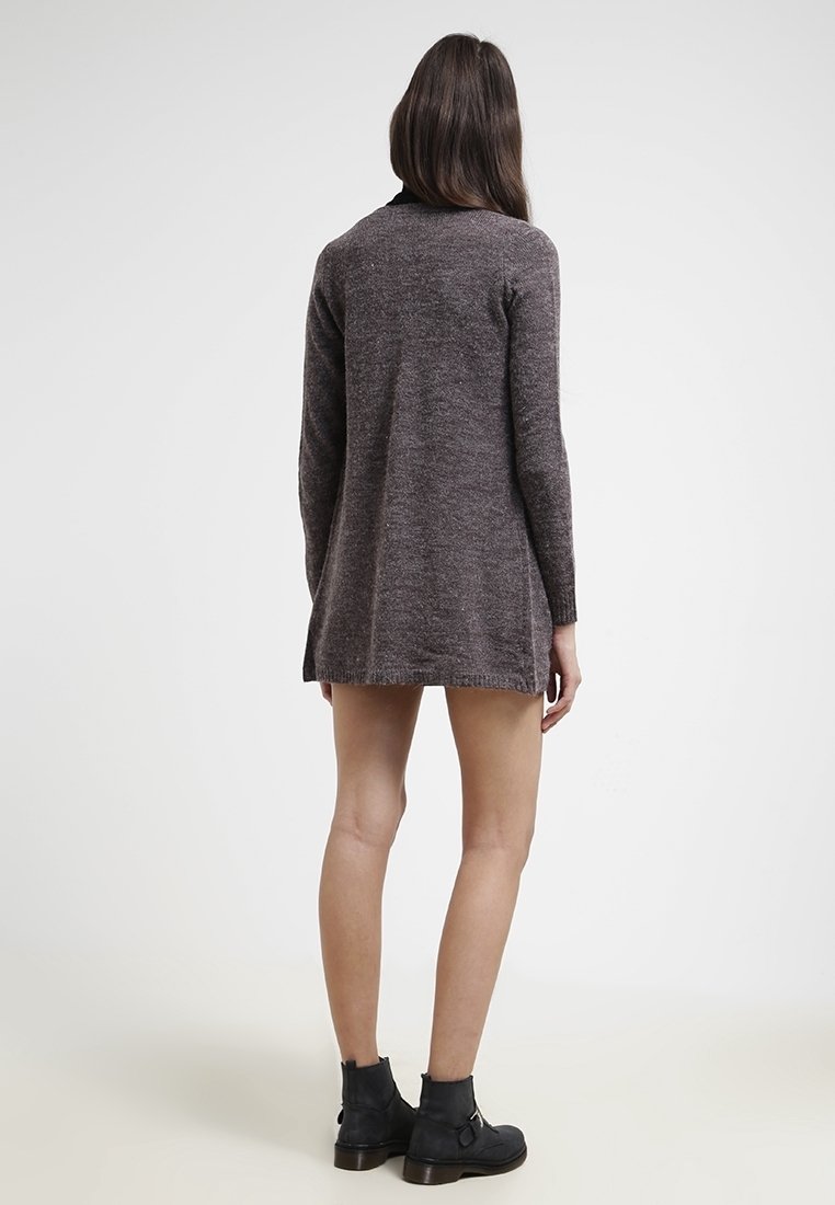 Femme debout de dos portant une robe-pull à manches longues marron foncé et des bottines noires sur un fond uni.