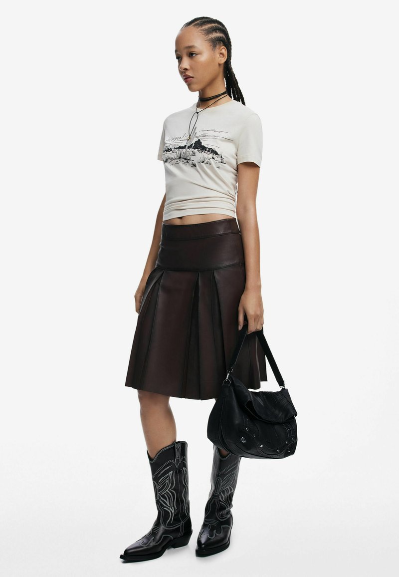 Hellbeige grafisches T-Shirt, brauner Lederfaltenrock, schwarze Cowboy-Stiefel mit weißer Naht und eine strukturierte schwarze Schultertasche.