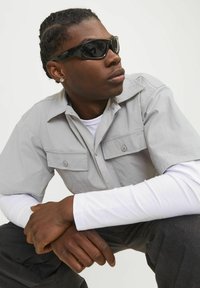 Schwarze Sonnenbrille, graues kurzärmliges Hemd mit zwei Brusttaschen, übereinander getragen mit einem weißen langärmligen Hemd und dunklen Hosen. Hände auf den Knien gefaltet.