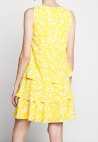 Žluté květinové šaty s bezramínkovým designem a volánky na lemu, vyrobené z lehké tkaniny s texturovaným povrchem.