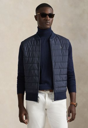 Granatowa pikowana kamizelka puchowa z stojącym kołnierzem, noszona na ciemnym golfie. Dopasowany fason z zamkiem błyskawicznym i ściągaczami przy mankietach.