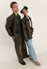 Két modell túlméretezett barna bőrdzsekiket, texturált zöld pulóvert, bő szárú sötétkék és világoskék farmert, valamint bézs papucsokat visel.