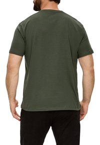 Atgalinė šono peržiūra trumpomis rankovėmis, alyvuogių žalia t-shirt pagaminta iš medvilnės. Yra apvali iškirptė ir laisvas siluetas. Dėvimos juodos kelnės.