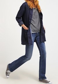 Veste longue bleu marine sur un haut gris à motifs, associée à un jean bleu et des baskets grises. La tenue présente une coupe décontractée et peu de détails.