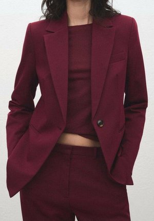 Kvinde iført en burgunderdragt med en enkeltknappet blazer, matchende bukser og en cropped top, hænderne i blazers lommer.