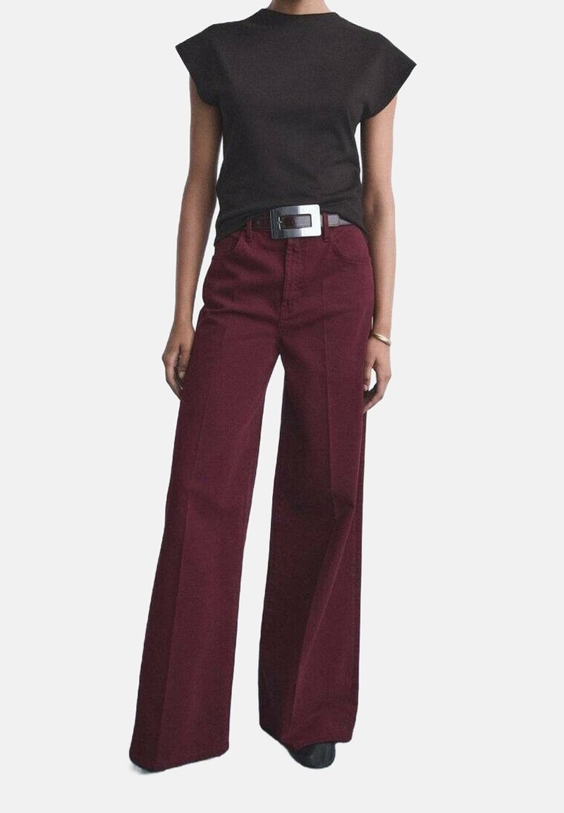 Personne portant un haut noir sans manches rentré dans un pantalon bordeaux taille haute à jambes larges, avec une ceinture noire avec une grande boucle rectangulaire.