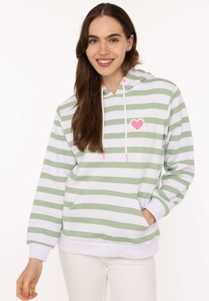 Junge Frau lächelt, trägt weißen und hellgrün gestreiften Hoodie mit pinkem Herzdetail und weiße Hose, Hand in der Tasche.