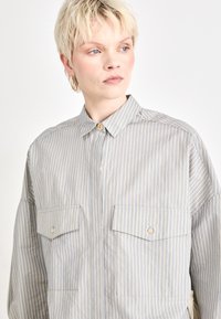Camicia a righe botton-up in blu chiaro e crema, dotata di due tasche sul petto, colletto e una vestibilità ampia con una texture morbida.