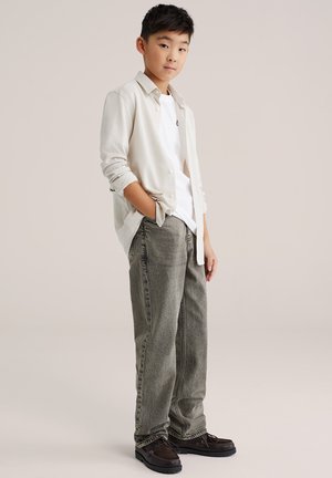 Vaaleanbeige neuletakki valkoisen T-paidan päällä, yhdistettynä löysiin harmaisiin farkkuihin ja tummanruskeisiin kenkiin. Yksinkertainen muotoilu, rento istuvuus.