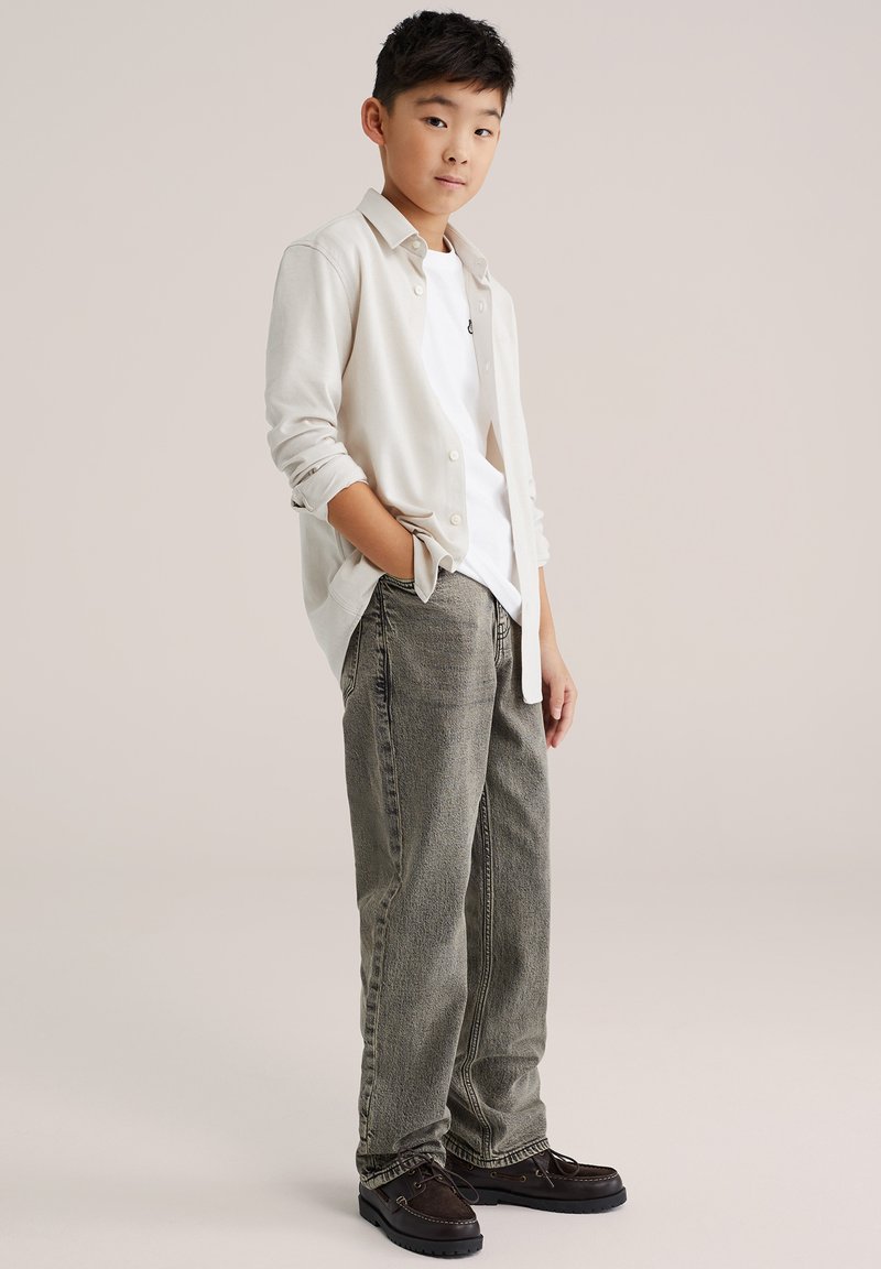 Világos bézs kardigán fehér póló felett, laza, szürke farmernadrággal és sötétbarna cipővel párosítva. Egyszerű dizájn, laza fazon.