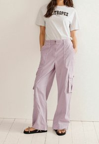 Pantalon cargo lilas avec poches latérales, présentant une coupe décontractée, en coton, avec passants pour ceinture et fermeture par bouton. Associé à un t-shirt graphique gris.