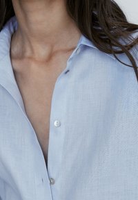 Camicia in lino azzurro chiaro con colletto e un leggero dettaglio con bottone, texture verticale e vestibilità rilassata.