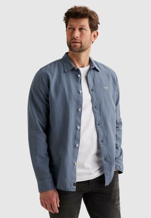 Homme portant une chemise bleue à manches longues déboutonnée par-dessus un t-shirt blanc et un jean foncé, regardant légèrement vers la droite sur un fond uni.