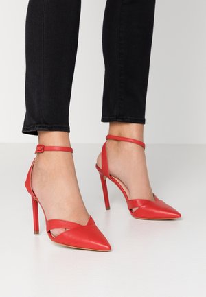 Pieds portant des escarpins rouges à bout pointu avec des brides à la cheville, assortis à un pantalon noir slim sur un fond uni.