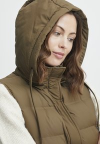 Brūns polsterēts veste ar kapuci, ar diegu detaļām, ar rāvējslēdzēja aizdari un gludu tekstūru. Akcentēta ar krēmkrāsas džemperi zem tās.