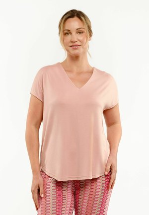 Kobieta o blond włosach ma na sobie jasnoróżową koszulkę z krótkim rękawem i dekoltem w serek oraz wzorzyste, różowe, czerwone i białe spodnie, stoi na białym tle.