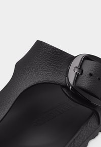 Κοντινό πλάνο λουριού μαύρου σανδαλιού με υφή στην επιφάνεια και γυαλιστερή μαύρη ρυθμιζόμενη αγκράφα σε απλό φόντο.