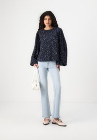 Blusa navy con motivo a pois, maniche lunghe a sbuffo, abbinata a jeans chiari a palazzo e scarpe beige slip-on; borsa bianca in mano.