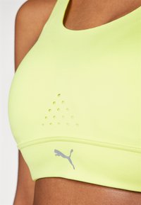 Sujetador deportivo de tejido amarillo brillante, con perforaciones tipo malla en un lado, logo gris en la parte inferior y una textura suave.