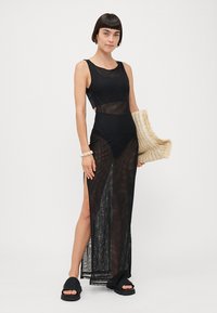 Abito maxi nero con pannelli in rete, spacco alto e corsetto aderente. Abbinato a sandali neri spessi e con in mano una borsa intrecciata.