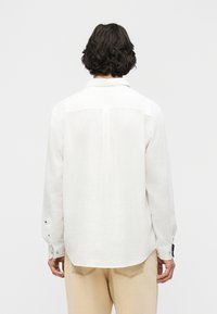 Άτομο με σκούρα, σγουρά μαλλιά μήκους μέχρι τους ώμους, που φοράει λευκό πουκάμισο με μακριά μανίκια και μπεζ παντελόνι, βλεπόμενο από πίσω.