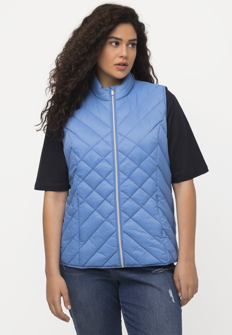 Gilet trapuntato azzurro chiaro con colletto alto e zip frontale. Abbinato a una maglietta nera a maniche corte e jeans in denim, con un ginocchio strappato.