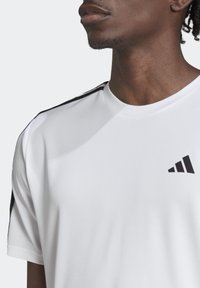 Lähellä kuvattuna henkilö, joka pitää yllään valkoista urheilupaitaa, jossa on mustat raidat olkapäillä ja pieni musta Adidas-logo rinnassa.